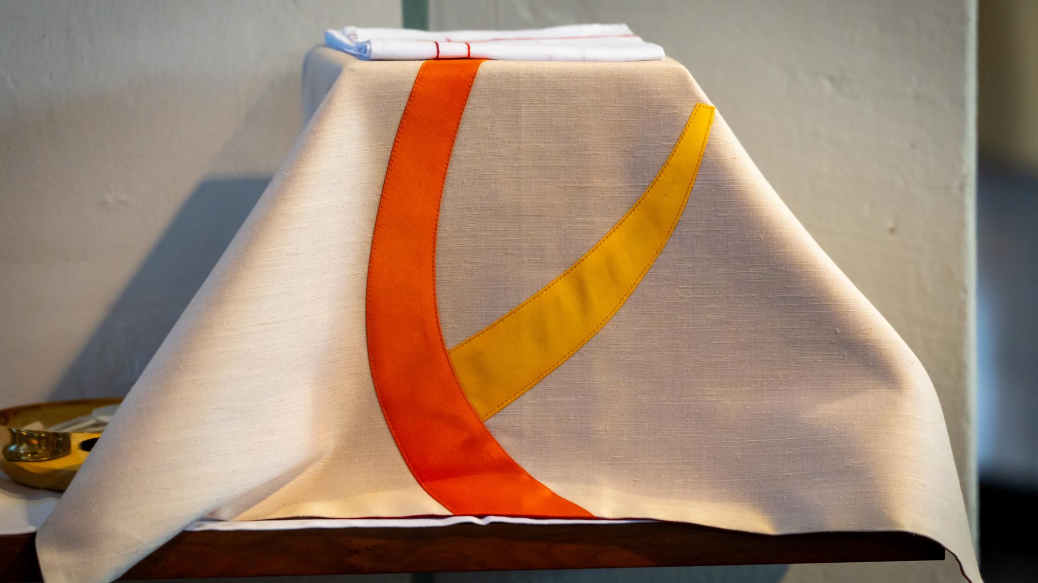 Ein helles Kelchvelum mit orange- und gelbfarbenen Verzierungen liegt sorgfältig auf einem Kelch. Die Aufnahme zeigt die Vorbereitung für einen festlichen Gottesdienst im Kirchenraum.