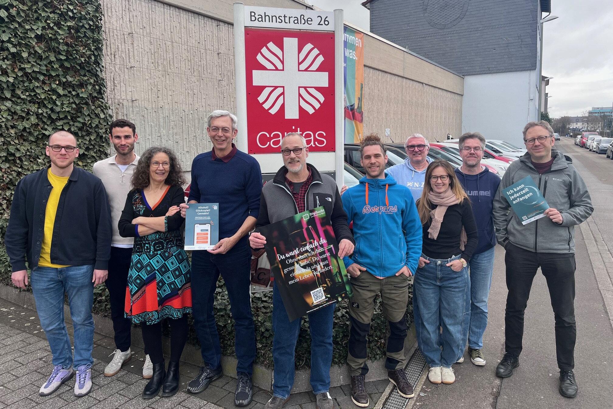 Eine Gruppe von unterschiedlichen Leuten steht draußen vor dem Caritas-Logo und schaut in die Kamera.